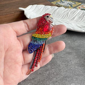 May include: A colorful parrot brooch with a red, orange, yellow, green, and blue crystal design. The parrot has a red heart-shaped crystal on its chest and a silver beak. The brooch is a decorative accessory.
