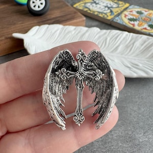 May include: A silver-colored pendant featuring a cross design with ornate detailing, flanked by detailed angel wings. The wings are spread out, creating a symmetrical design. The pendant is held in a hand.