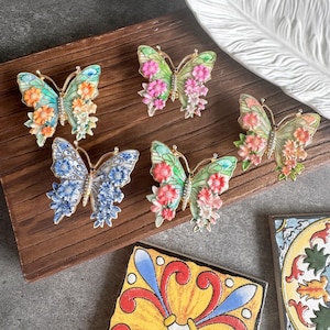 May include: Five butterfly-shaped brooches with gold-tone metal bodies and wings decorated with colorful enamel floral patterns. Each butterfly features a central row of small clear stones along the body, with wings displaying various floral designs in shades of blue, green, pink, and orange.