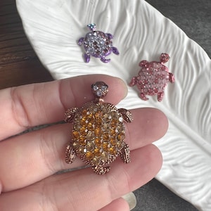 May include: Three turtle-shaped brooches featuring metallic bodies encrusted with small, sparkling rhinestones. One brooch is amber-coloured, one is pink, and one is purple. Each turtle has a small head, four legs, and a detailed shell design.