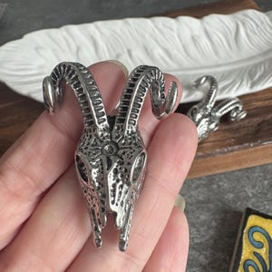 May include: A silver-toned metal pendant shaped like a ram skull with curved horns and a textured surface. The skull features a star-like symbol etched on the forehead area between the horns. The piece has a metallic finish and intricate detailing.