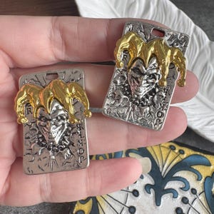Op de afbeelding: Twee zilverkleurige hangers met een goudkleurig narrenhoedontwerp. Elke hanger heeft een lachend narrengezicht en het woord "JOKER" op een rechthoekige kaart. De hangers hebben een vintage uitstraling.