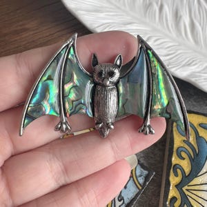May include: A silver bat brooch with iridescent wings. The bat's wings have a shimmering display of greens and blues. The brooch features a textured body and small, pointed ears, ideal for adding a touch of gothic flair to any outfit.