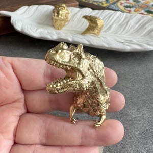 Può includere: Una dettagliata statuetta dorata di Tyrannosaurus Rex con la bocca aperta, che mostra i denti. Il dinosauro è tenuto in mano, con altri oggetti decorativi dorati sullo sfondo su un piatto bianco a forma di piuma.