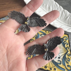 May include: Two detailed, dark metal eagle figurines with outstretched wings. The eagles have gold accents on their chests and beaks. The figurines are displayed on a hand, showcasing their intricate design and craftsmanship.