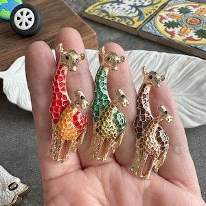 May include: Three gold-toned giraffe brooches with colorful enamel detailing. One brooch features red and orange, another green and yellow, and the third brown and gold. Each brooch has a baby giraffe.