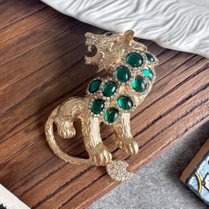 Op de afbeelding: Een goudkleurige metalen broche in de vorm van een brullende tijger. Het lichaam is getextureerd om op vacht te lijken en is versierd met verschillende ovale, donkergroene cabochonstenen, omringd door kleine, heldere strass-steentjes langs de nek.