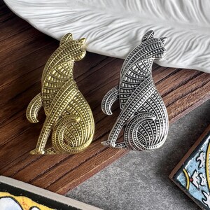 Op de afbeelding: Twee katvormige broches, één in een goudkleurige afwerking en één in een zilverkleurige afwerking. Elke broche heeft een gedetailleerd, gestructureerd patroon van lijnen en geometrische vormen over het lichaam, met de kat zittend en de staart gekruld.