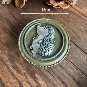 Pode incluir: Um botão redondo de metal em estilo antigo com um design de ouriço prateado. O botão tem 3,5 cm de diâmetro e tem uma aparência detalhada e texturizada. O botão tem uma borda de cor bronze e um centro de tom verde.
