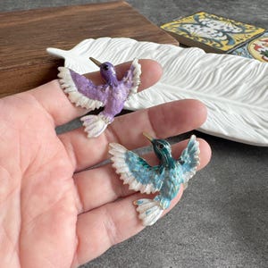 May include: Two hummingbird brooches with outstretched wings. One is purple with white accents, the other is teal with white accents. Both have gold-colored beaks and are set against a neutral background.