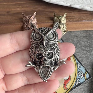 May include: A silver-toned metal brooch shaped like an owl with a skull integrated into its chest, perched on a branch. The design features intricate engraved details on the feathers and eyes, with a metallic finish and a three-dimensional structure.