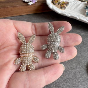 May include: Two jeweled bunny figurines. One is gold-toned, the other silver-toned. Both are encrusted with small, sparkling crystals and have pearl accents around the neck. The bunnies have long ears and small arms and legs.