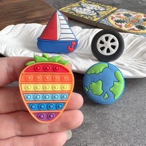 Op de afbeelding: Een verzameling kleurrijke bedels, waaronder een rood, wit en blauw zeilbootje, een zwarte band met een witte rand, een regenboogkleurige aardbei en een blauwe en groene wereldbol. De bedels liggen op een wit, golvend oppervlak.