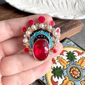 May include: A gold-tone brooch featuring a large oval red faceted center stone, surrounded by smaller red enamel circles, white faux pearls, and clear rhinestones. The design includes black and turquoise enamel accents in a fan-like shape.