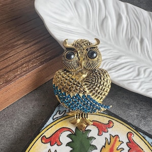 May include: A gold-toned owl brooch featuring two prominent black circular eyes and a textured feather pattern across the body. The lower portion of the owl's wings is embellished with a dense arrangement of small, vibrant blue rhinestones.