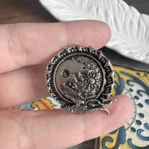 May include: A circular silver-toned metal pin featuring a textured relief design of a wolf howling at a sun or moon. The outer edge of the pin has a jagged, spiked border, and the central surface has a dark, weathered finish with raised details.