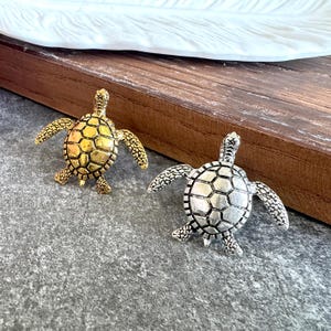 May include: Two turtle-shaped decorative pins, one finished in a polished gold tone and the other in a brushed silver tone. Each turtle features a detailed, textured shell with a geometric pattern and four outstretched flippers, crafted in metal.