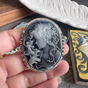 May include: An oval brooch featuring a black and white cameo relief of a figure with flowing hair adorned with flowers, holding a small winged creature. The frame is silver-toned metal with a row of small, dark, circular inset stones along the edge.