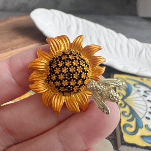 Peut inclure: Une broche tournesol avec des pétales jaune doré et un centre brun foncé aux détails en relief. Une abeille métallique argentée, dotée d'ailes et d'un corps texturés, est fixée sur le côté droit de la fleur, reposant sur les pétales.