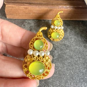 Può includere: Un paio di orecchini in metallo dorato a forma di zucca, caratterizzati da un intricato lavoro in filigrana, piccole perle bianche e pietre cabochon traslucide color verde lime incastonate al centro di ogni pezzo decorativo.