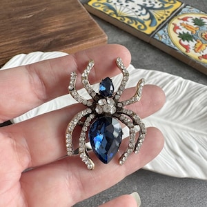 May include: A gold-toned spider brooch with clear crystal accents and two large, deep blue teardrop-shaped gemstones. The spider's legs and body are adorned with sparkling crystals, creating a glamorous accessory.