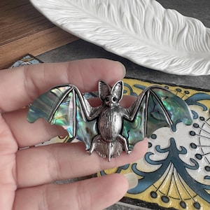 May include: A silver-toned bat-shaped brooch with iridescent abalone wings. The bat has detailed features, including small ears and eyes. The wings display shades of green, blue, and purple. The brooch is held in a hand.