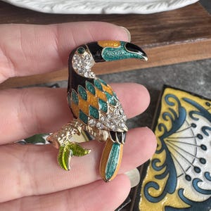 May include: A toucan brooch featuring gold-tone metal, black, teal, and yellow enamel in a diamond pattern, accented with clear rhinestones along the neck and wing. The bird is perched on a textured branch with a small green leaf at the base.