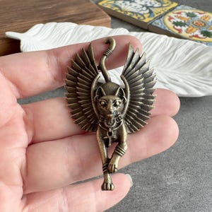 May include: Bronze cat pendant with wings, a stylized tail, and an ankh pendant. The cat is in a walking pose, with detailed wings and body. The pendant has an antique finish, with a vintage aesthetic.