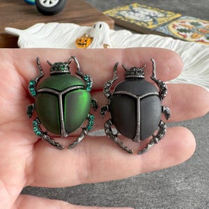 Peut inclure: Deux broches en forme de scarabée, une verte et une noire, tenues dans une main. Les deux ont des détails en métal argenté et des accents de bijoux. La broche verte a une finition métallique. Accessoires de mode.