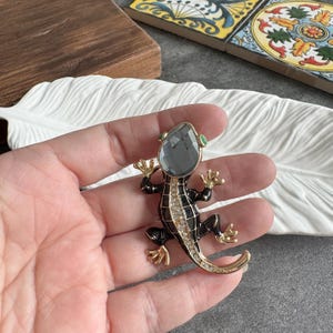 Op de afbeelding: Een gedetailleerde broche in de vorm van een hagedis. De hagedis heeft een zwart en goudkleurig lichaam met een grijze edelsteen kop en kleine groene accenten. De broche is versierd met heldere kristallen.