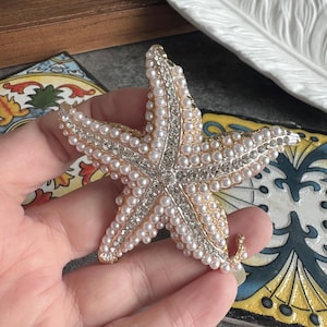 May include: A starfish-shaped brooch featuring a gold-tone metal base encrusted with numerous small white faux pearls and a central strip of clear sparkling rhinestones running along each of the five arms, meeting at the centre point.