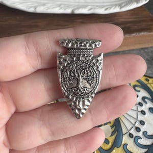 May include: A silver-toned metal pendant shaped like an arrowhead with a textured, hammered surface. The centre features a circular embossed design of a tree with intricate branches and roots, framed by a rope-like border at the top section.