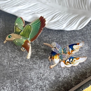Könnte beinhalten: Zwei Kolibri-Broschen. Die linke Brosche zeigt grüne, braune und goldene Emaille mit einem einzelnen grünen Stein als Auge. Die rechte Brosche zeigt blaue, weiße und orangefarbene Emaille mit violetten, türkisfarbenen und klaren Kristallakzenten.