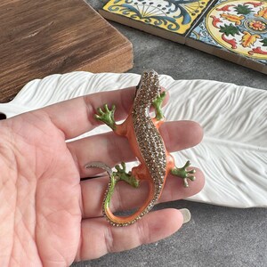 Könnte beinhalten: Dekorative Eidechsenbrosche mit orangefarbenem und grünem Körper, verziert mit funkelnden Strasssteinen. Der Schwanz der Eidechse ist silberfarben. Die Brosche wird in einer Hand gehalten und liegt auf einer weißen, federförmigen Schale.