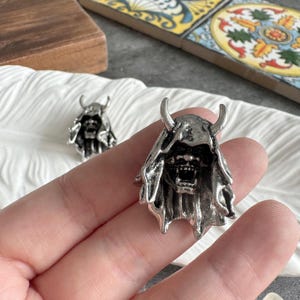 Può includere: Primo piano di due ciondoli a forma di maschera Oni color argento. I ciondoli presentano un design dettagliato di un demone cornuto con bocca aperta e cappuccio. I ciondoli sono su una superficie bianca strutturata.