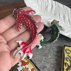 Op de afbeelding: Twee koi-vis broches met een metallic emaille afwerking. De ene broche is rood met witte accenten op de staart en rode kristallen op de kop. De tweede broche is groen met heldere kristallen op de kop. Beide hebben zilverkleurige metalen randen.