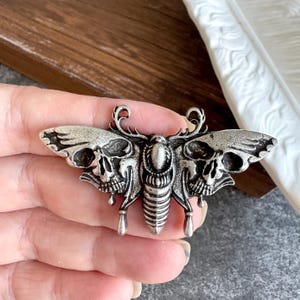 May include: A silver-toned metal brooch shaped like a moth with intricate details. The wings feature prominent skull patterns on each side, while the central body is segmented. The design includes curved antennae at the top and two small dangling elements.