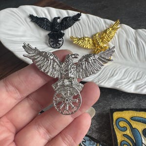 May include: Silver, gold, and black eagle brooches with outstretched wings and a wheel design. The brooches are displayed on a white, feather-shaped dish. The brooches are detailed and have a metallic finish. The brooches are likely for decorative purposes.