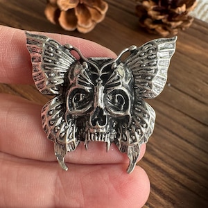 Pode incluir: Um broche de metal prateado em forma de borboleta com um design central de caveira. As asas apresentam padrões gravados intrincados e detalhes escuros em relevo, enquanto a caveira forma o corpo da borboleta com órbitas e dentes visíveis.