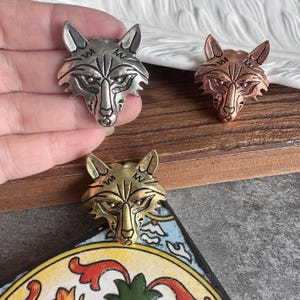 Puede incluir: Tres dijes de metal con forma de cabeza de lobo en tonos plata, cobre y oro. Cada dije presenta líneas grabadas detalladas que definen las orejas, los ojos y el hocico del lobo. El dije de color plata se sostiene con la mano.