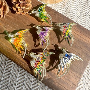 Op de afbeelding: Zes vogelvormige broches met metalen lichamen bezet met heldere strass-steentjes en kleurrijke emaille vleugels. Elke broche heeft een centrale ovale edelsteen in kleuren zoals groen, paars, rood, geel, oranje en blauw.