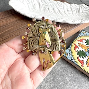 Può includere: Una spilla in metallo dorato a forma di leone stilizzato con una grande criniera testurizzata. Il leone presenta occhi verdi, una lingua rosa e un corpo decorato con piccoli strass rotondi multicolori nelle tonalità ambra, marrone e trasparente.