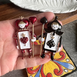 May include: Two enamel pins shaped like playing card characters. The left pin features a red heart suit with a crown and heart staff. The right pin features a black spade suit with a crown and spade staff. Both have white rectangular bodies.