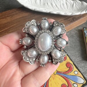 May include: A silver-toned brooch featuring a large central oval faux pearl surrounded by eight smaller oval faux pearls. The metal frame is embellished with numerous small, clear, sparkling rhinestones arranged in a floral-like pattern.
