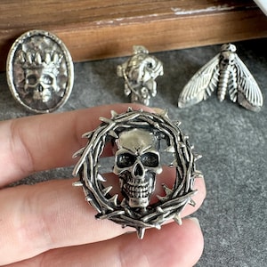 Può includere: Una spilla in metallo color argento raffigurante un teschio umano al centro di una corona di spine circolare. La spilla presenta una finitura metallica anticata con incisioni dettagliate sul teschio e sulle spine appuntite circostanti.