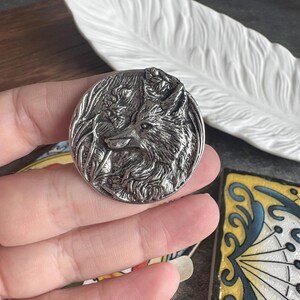 May include: A round, silver-toned pendant featuring a detailed relief of a wolf's head surrounded by foliage. The pendant is held in a hand, with a white feather-shaped dish and colorful tiles in the background.