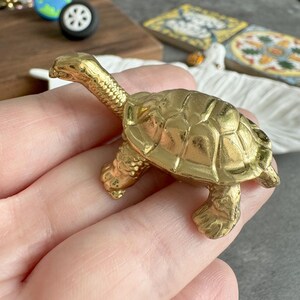 Könnte beinhalten: Kleine, goldfarbene Schildkrötenfigur. Die Schildkröte hat einen detaillierten Panzer, Kopf und Beine. Die Figur besteht aus einem glänzenden, metallischen Material. Die Schildkröte ist ein dekorativer Gegenstand.