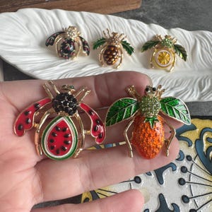 Peut inclure: Cinq broches en forme d'insectes avec des motifs de fruits et légumes. Deux sont tenues dans une main : l'une en forme de tranche de pastèque avec émail rouge et graines noires, l'autre en forme de fraise orange avec des cristaux verts.