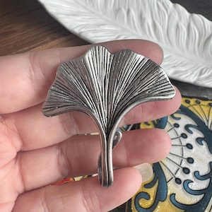 Puede incluir: Un broche de metal plateado con forma de hoja de ginkgo. La pieza presenta una silueta en forma de abanico con prominentes nervaduras verticales en relieve que irradian desde la base hasta el borde exterior, creando una superficie metálica texturizada.