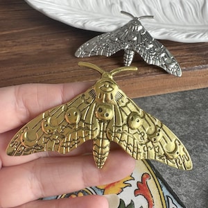 May include: A gold-toned metal moth brooch featuring embossed celestial details including crescent moons and circular patterns on the wings. A second silver-toned moth brooch with identical embossed patterns is positioned in the background.